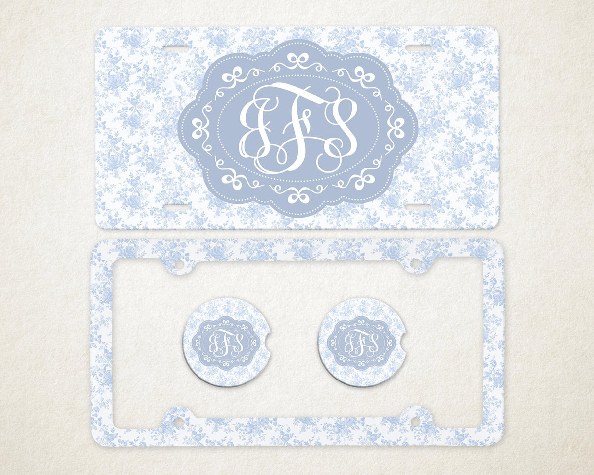 May include: A light blue license plate and frame set with a floral pattern. The license plate features a light blue oval with white initials. Two matching car coasters are also included.