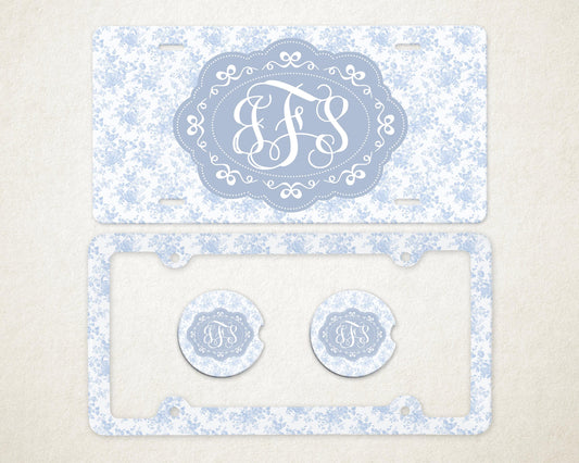 May include: A light blue license plate and frame set with a floral pattern. The license plate features a light blue oval with white initials. Two matching car coasters are also included.
