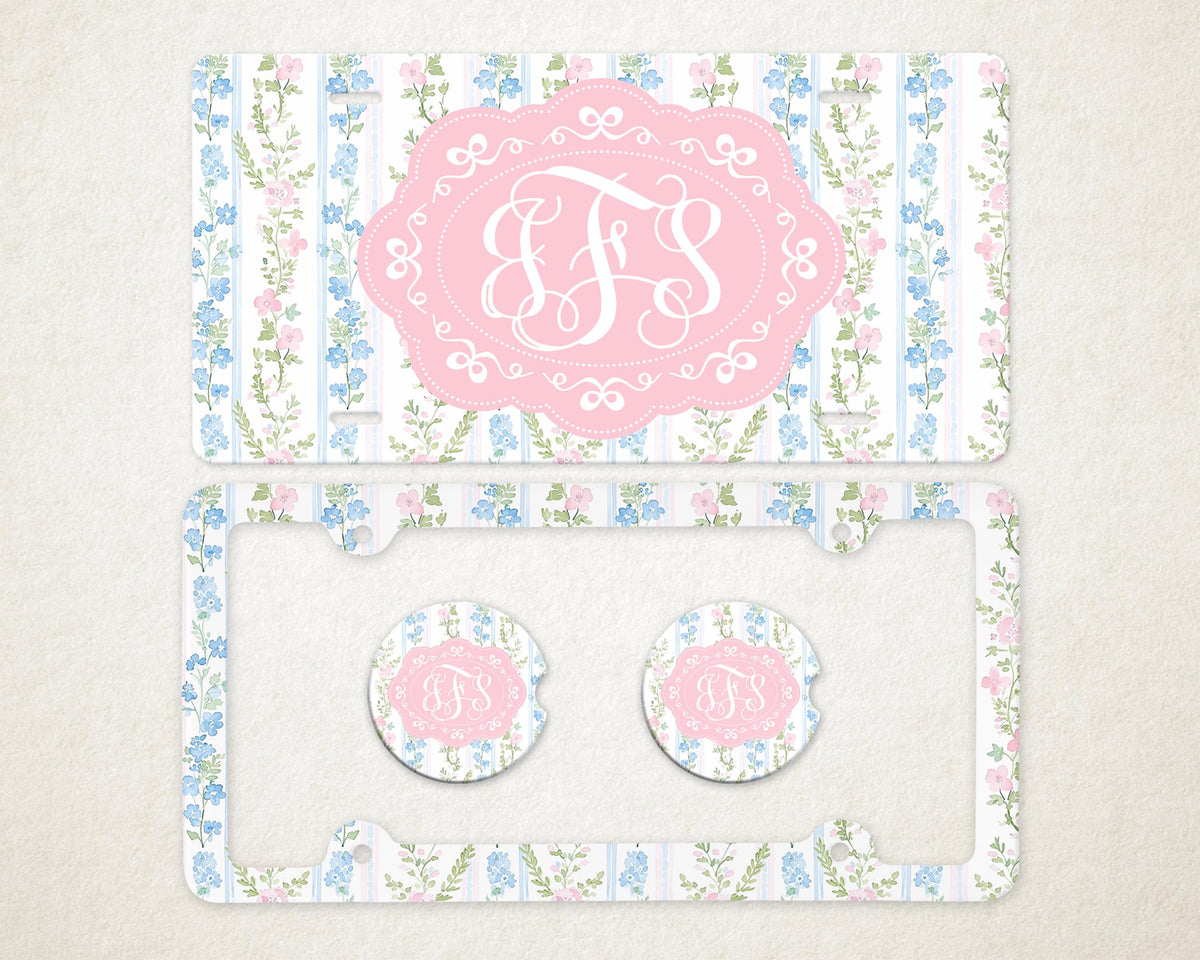 May include: A license plate, license plate frame, and two car coasters with a floral design. The items feature a pink and white monogram in a pink oval frame, set against a background of blue and pink flowers.