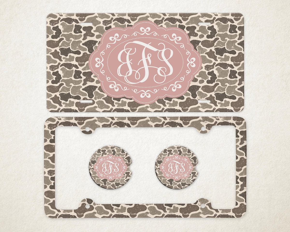 May include: A license plate, license plate frame, and two car coasters with a camouflage pattern in shades of brown and gray. Each item features a pink oval with a white monogram.