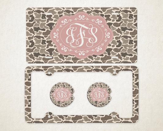 May include: A license plate, license plate frame, and two car coasters with a camouflage pattern in shades of brown and gray. Each item features a pink oval with a white monogram.
