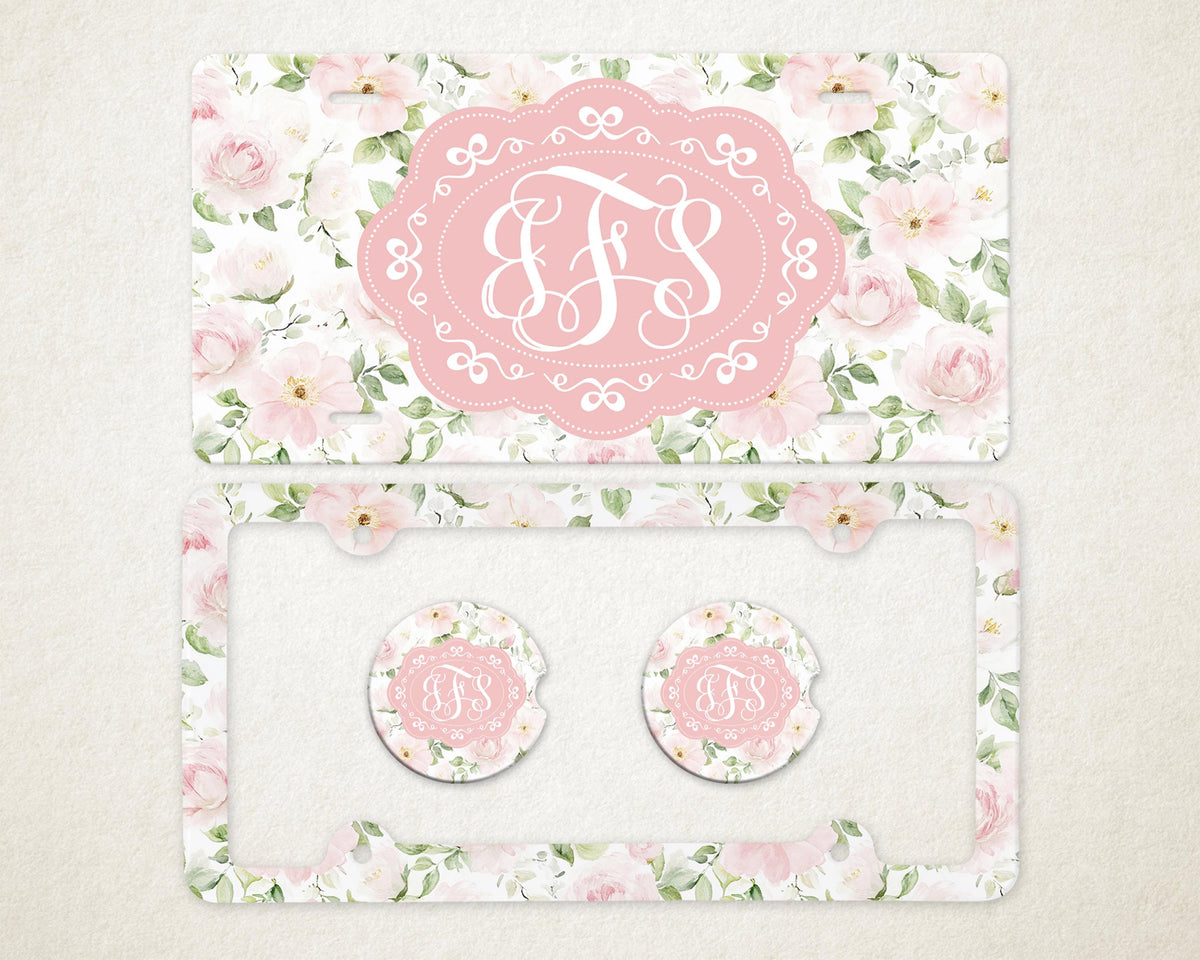 May include: A license plate and car accessories set featuring a floral design with pink roses and green leaves. The set includes a license plate, a license plate frame, and two car coasters. Each item has a pink oval with white initials.