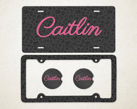 May include: A license plate and car coasters set. The license plate and coasters have a black leopard print design with the name "Caitlin" in pink script. The license plate frame is black.