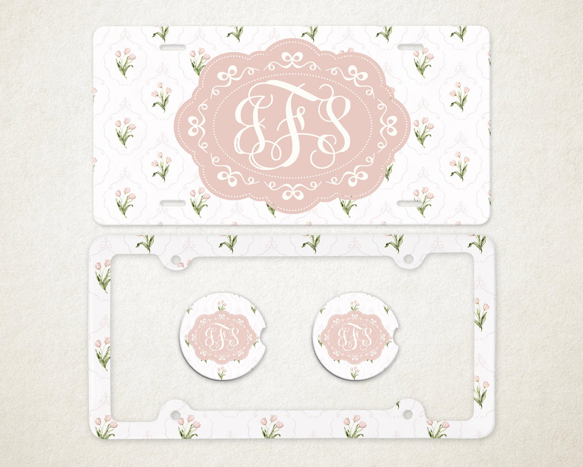 May include: A license plate, license plate frame, and two car coasters with a floral pattern and a monogram. The license plate and coasters have a pink oval with a white monogram. The frame and plate have a white background with pink flowers.