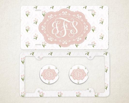 May include: A license plate, license plate frame, and two car coasters with a floral pattern and a monogram. The license plate and coasters have a pink oval with a white monogram. The frame and plate have a white background with pink flowers.
