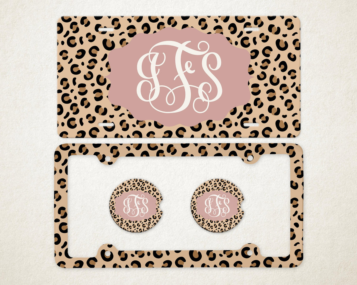 May include: A license plate and frame set with matching car coasters. The set features a tan leopard print background with brown spots. A pink oval shape in the center of the license plate and coasters displays a white monogram. The license plate frame is also leopard print.