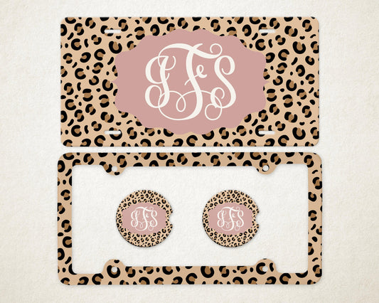 May include: A license plate and frame set with matching car coasters. The set features a tan leopard print background with brown spots. A pink oval shape in the center of the license plate and coasters displays a white monogram. The license plate frame is also leopard print.